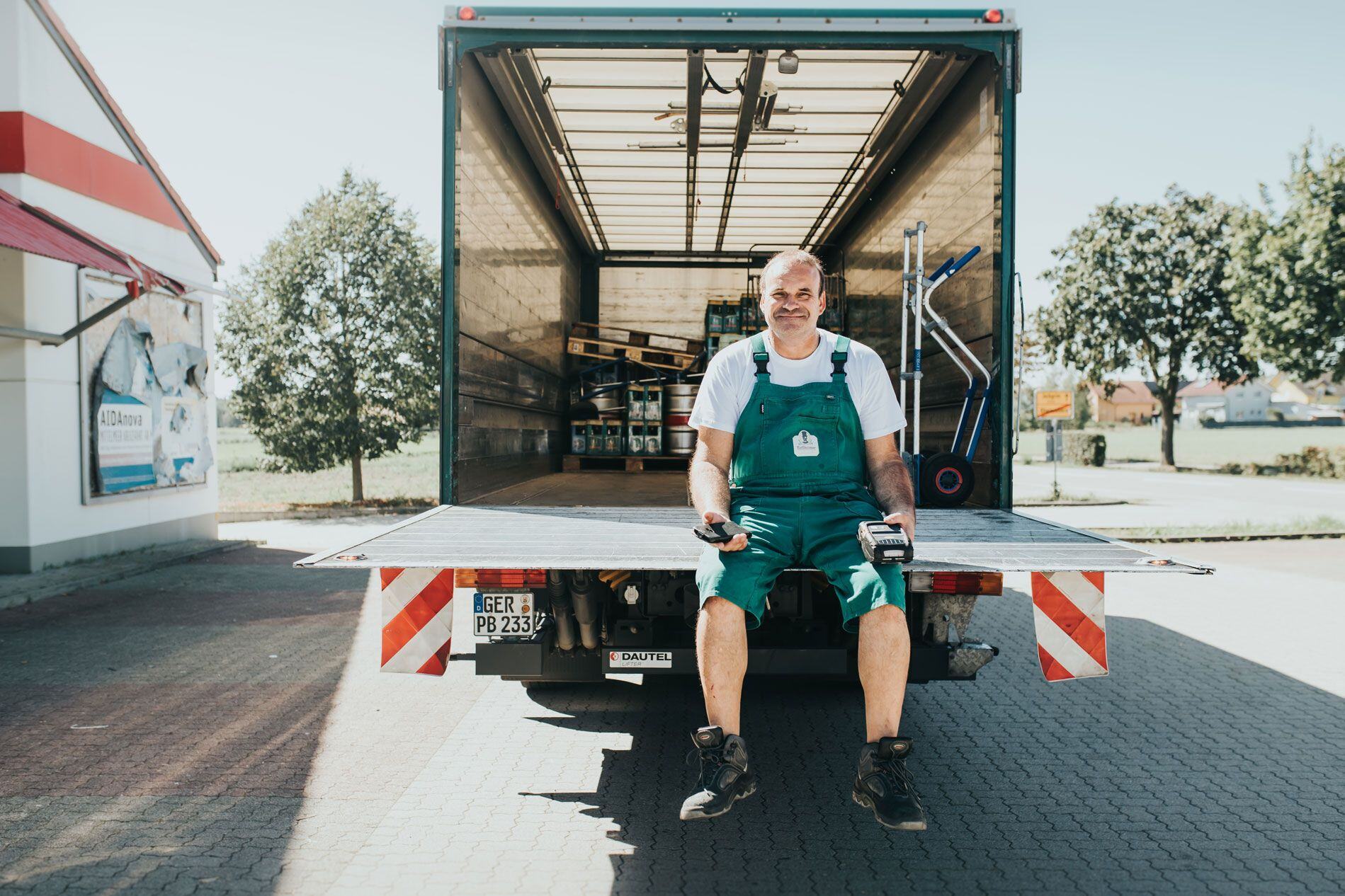 A delivery worker in green overalls sits on the tailgate of a truck, smiling and holding handheld devices.